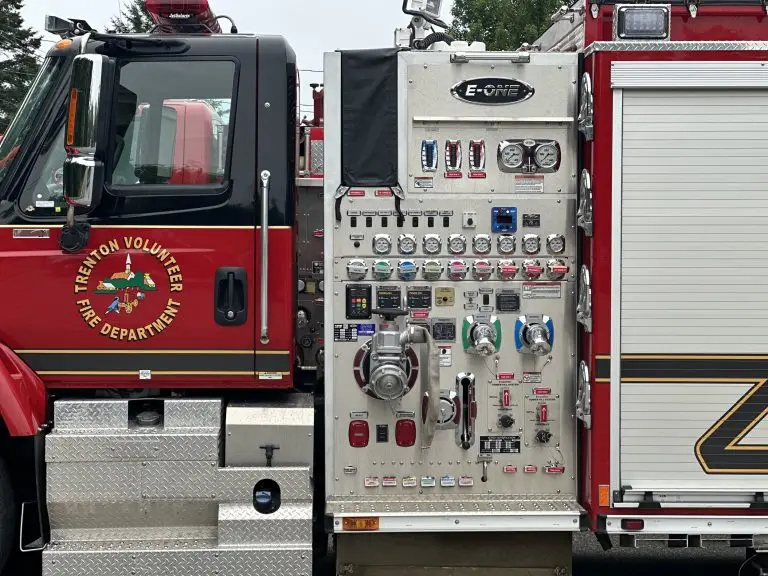A red fire engine from the Trenton Volunteer Fire Department is shown close-up. The side panel displays numerous gauges, dials, switches, and control handles used for operating the firefighting equipment. The fire department's emblem is visible on the door.