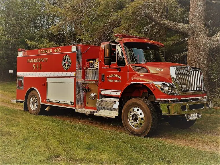 A red fire truck labeled "Tanker 402" and "Lamoine Fire Dept." is parked on a grassy area beside a large tree. The truck features emergency contact information and the International Harvester logo on its front grille.