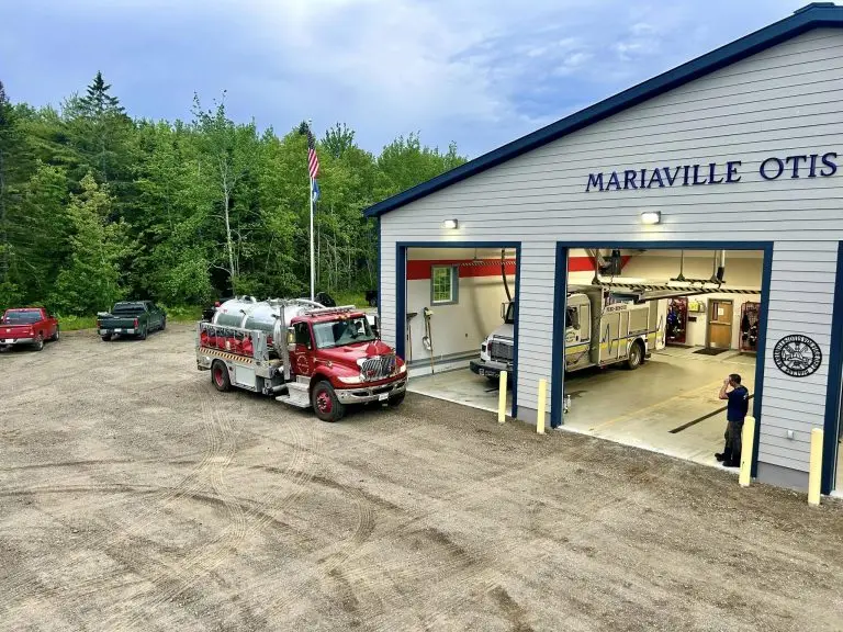 A fire station with the sign "Mariaville Otis" has its garage door open, revealing a fire truck inside. Another fire truck is parked outside, next to a person on the right side of the image. Several vehicles are parked on the gravel driveway near the station. Trees are in the background.