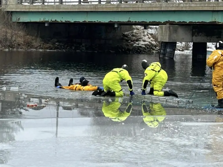 Three rescue workers in yellow protective suits assist a person lying on an icy surface near a bridge. The workers appear to be securing and helping the person, possibly after an accident on the ice. The scene reflects the urgency of a rescue operation.