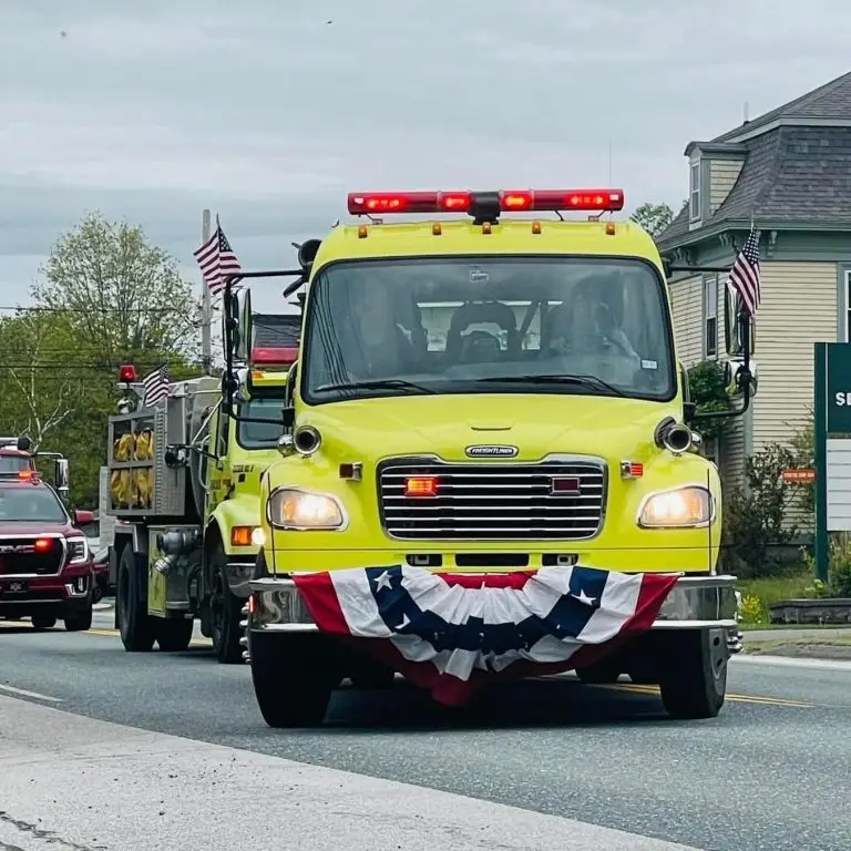 A yellow fire truck adorned with American flags and a red, white, and blue banner drives down a street, participating in a parade. Another emergency vehicle is visible behind it. Houses and trees line the street under a cloudy sky.